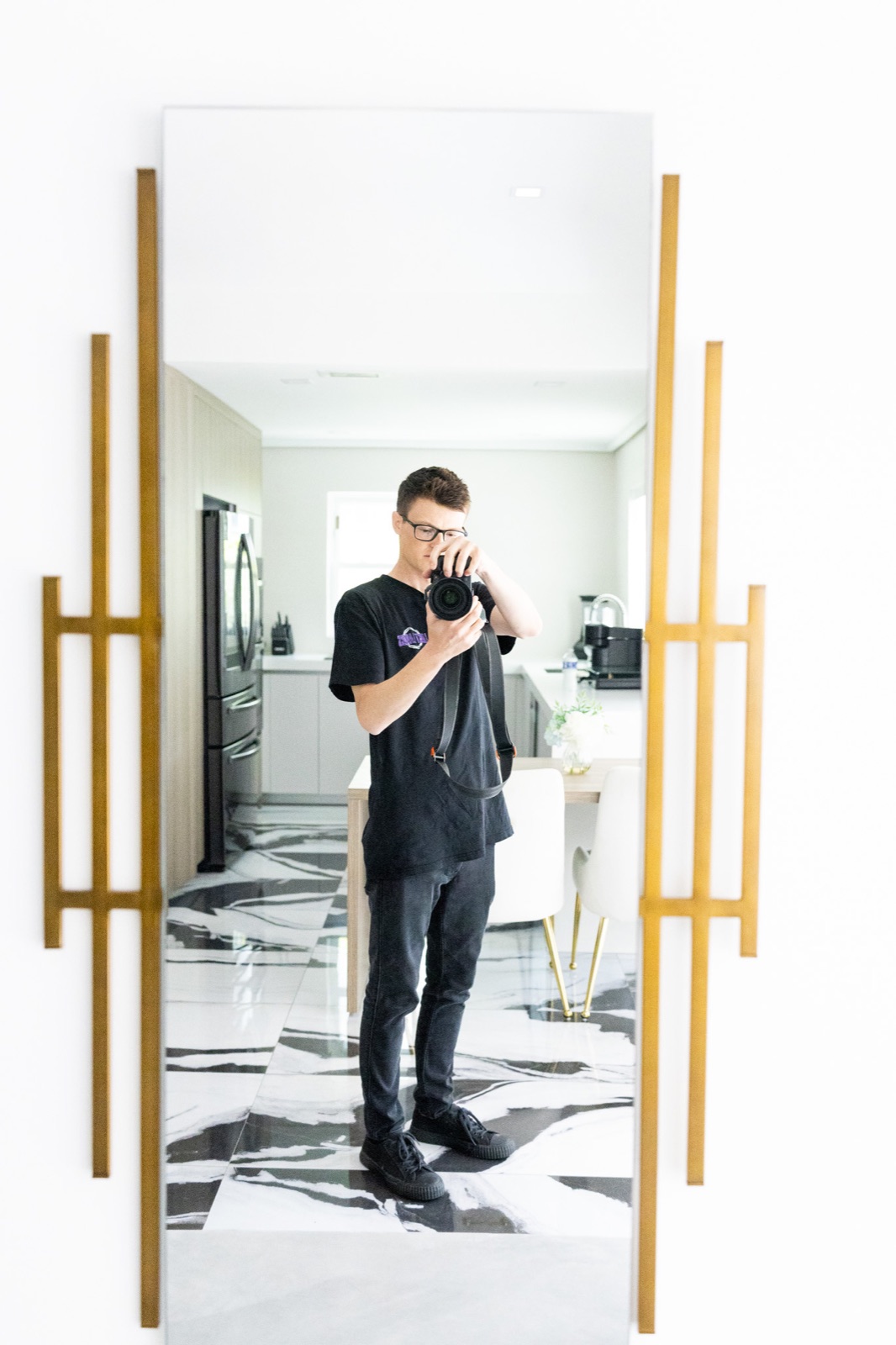 A person wearing glasses holds a camera in a tall gold-framed mirror in a bright room.
